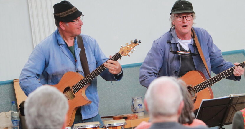 Franz Offenbecher und Klaus Lohr (v.li.) bei einem früheren Auftritt in der TV Halle Erfelden. Franz Offenbecher und Klaus (v.li.) stehen mit ihren Gitarren vor ihrem Publikum. Von den Zuhörerinnen und Zuhörern sind einige Hinterköpfe zu sehen.