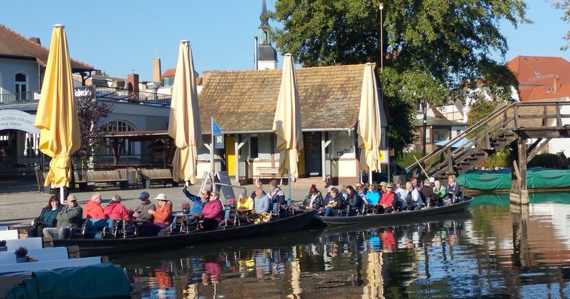 Auch Bootsfahrten im Spreewald stehen auf dem Programm. Mehrere lange Kanus haben längs an einer Uferpromenade festgemacht. In den Booten sitzen viele Menschen.