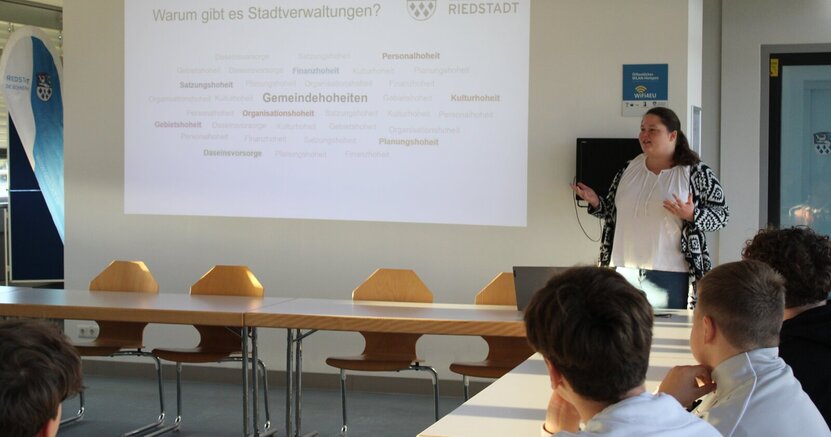 Julia Galley gab einen Überblick über die Aufgaben einer Stadtverwaltung. Julia Galley steht vor einer Wand im Sitzungssaal. Neben ihr wird eine Präsentation zu den Aufgaben der Stadtverwaltung an der Wand abgebildet.