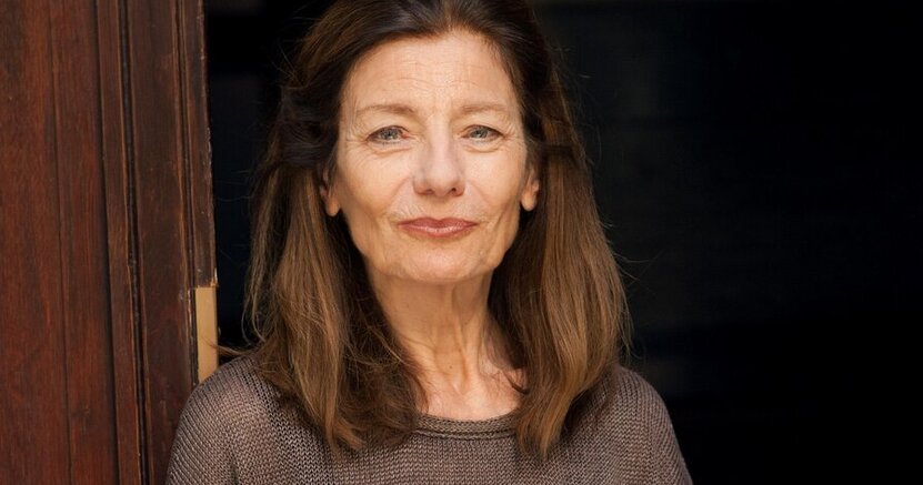 Ursula Krechel kommt nach Riedstadt. Ursula Krechel hat ein braunes Oberteil mit kurzen Ärmeln an und lehnt sich an einen dunkelbraunen Türrahmen.