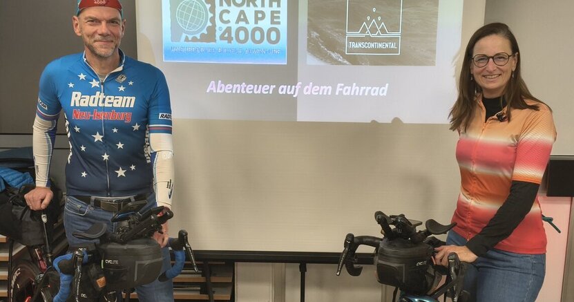 Tom und Stevie Thissen freuen sich auf ihren Multimedia-Vortrag im Alten Rathaus Crumstadt. Tom und Stevie Thissen stehen mit ihren Fahrrädern im Saal des alten Rathauses Crumstadt. Zwischen ihnen befindet sich eine Leinwand. Drauf ist die Ankündigung ihres Vortrags zu lesen.