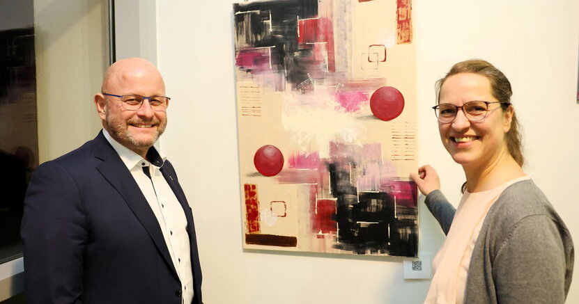 Bürgermeister Marcus Kretschmann und Künstlerin Aida Hiseni vor einem Werk der Künstlerin. Bürgermeister Marcus Kretschmann und Künstlerin Aida Hiseni stehen links und rechts vor einem Werk der Künstlerin. Es ist ein abstraktes Gemälde mit bunten Kugeln und rechteckigen Formen.