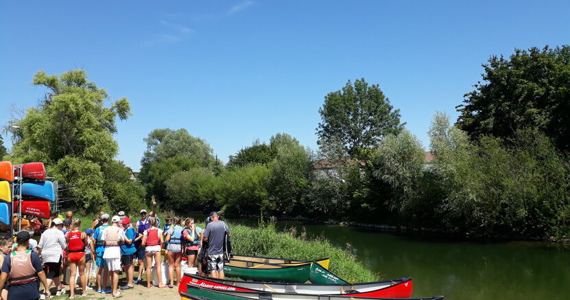 Auch eine Kanutour gehört zum Programm der Teeniefreizeit Links von einem Fluss liegen auf dem Ufer mehrere Kanus. Jugendliche in Schwimmwesten stehen daneben, mit dem Rücken zum Betrachter.