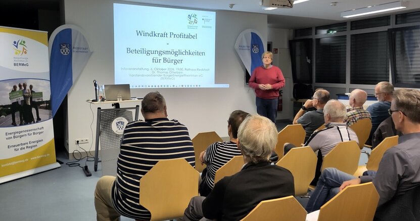 Referent Thomas Otterbein erklärt dem interessierten Publikum die BERMeG Von hinten sind vollbesetzte Stuhlreihen im Sitzungssaal des Rathauses zu sehen. Vorne steht ein Mann im roten Pullover neben einer an der Wand gezeigten Präsentation. Darauf steht gerade "Windkraft profitabel. Beteiligungsmöglichkeiten für Bürger.