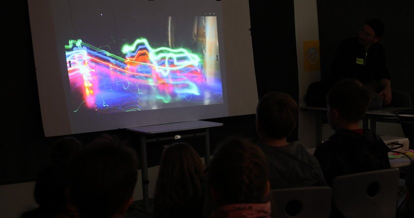 Beim "Winter-Fun" gab es bereits einen ersten Kunstworkshop, der sich mit verschiedenen Foto- und Maltechniken beschäftigte. Kinder sitzen vor einer Leinwand und schauen sich eine Fotografie an, die aus bunten Lichtstreifen besteht.