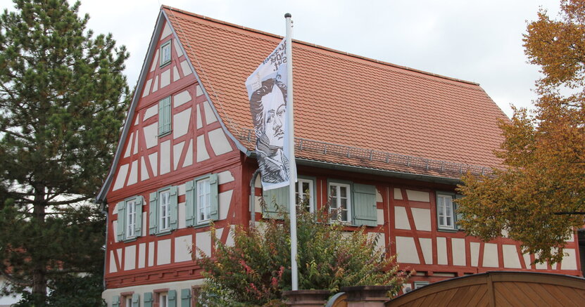 Das Büchnerhaus in Riedstadt-Goddelau. Zu sehen ist die Frontseite des Büchnerhauses, ein altes Fachwerkhaus mit roten Balken. Vor dem Haus weht eine Fahne mit dem gezeichneten Porträt Georg Büchners.