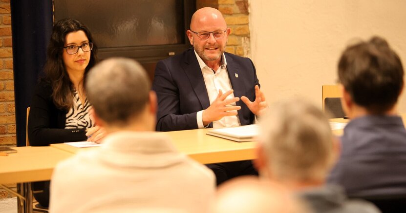 Bürgermeister Marcus Kretschmann mit Giulia Reinhardt vom Bürgerservice beim Bürgergespräch in Wolfskehlen. Giulia Reinhardt vom Bürgerservice und Bürgermeister Marcus Kretschmann sitzen an einem Tisch vor Publikum auf Stühlen. Von den Zuhörern sind die Hinterköpfe zu sehen.