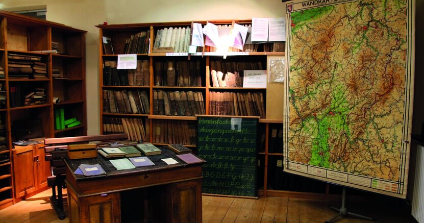 Im Heimatmuseum Crumstadt ist auch ein altes Klassenzimmer zu sehen. Zu sehen ist ein altes Klassenzimmer: Im Vodergrund steht ein altes Pult mit alten Heften und Büchern. An der Wand befinden sich alte Holzschränke mit Büchern. Rechts im Hintergrund hängt eine alte Wandkarte von Hessen.