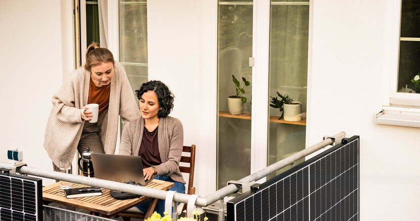 Mit PV und Köpfchen zur leistbaren Energiewende. Eine Frau sitzt auf ihrem Balkon an ihrem Laptop. Eine andere Frau steht neben ihr , hält eine Kaffeetasse in der Hand und schaut auf den Bildschirm des Laptops- An der Balkonumrandung ist eine PVC-Anlage angebracht.