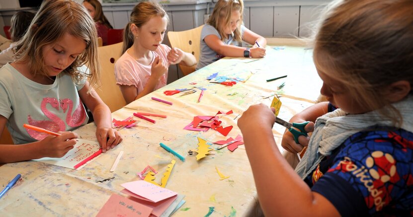 Basteln wie hier 2025 in Leeheim gehört auch zu den Angeboten der Ferienspiele. Kinder sitzen an beiden Seiten eines langen Tisches und malen und schneiden aus.
