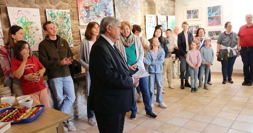 Erster Stadtrat Richard Kraft sprach zur Eröffnung der Kinderkunst-Ausstellung in der Kunstgalerie am Büchnerhaus. Erster Stadtrat Richard Kraft steht im Anzug in der Kunstgalerie am Büchnerhaus. An der Wand sind bunte Kindergemälde zu sehen. Davor stehen Eltern mit ihren Kindern und hören Kraft zu. An der Seite steht ein Tisch mit Kleinigkeiten zum Essen.