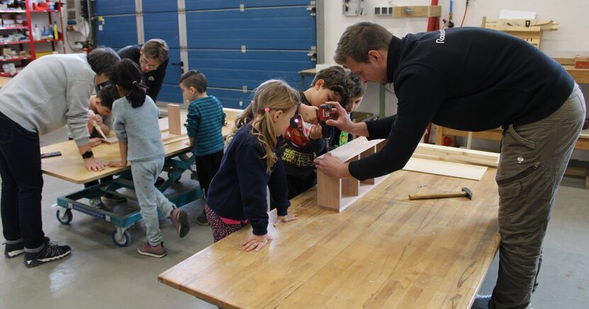 Anabelle, Emilio und Eymen (v.li.) bauen mit André Lindemann einen Kasten des Insektenhotels. In der Schreinerei stehen ein Mädchen und zwei Jungs auf einer Seite einer Arbeitsplatte. Auf der anderen Seite steht André Lindemann. Er hilft dem Jungen in der Mitte, mit einem Akkubohrer eine Schraube in einen Holzkasten zu schrauben.