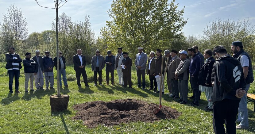 Mitglieder der Ahmadiyya-Muslim-Gemeinde sowie Bürgermeister Marcus Kretschmann (6. v.l.) und Karlheinz Hebermehl (5. v.l.) während der Ansprache von Rafique Khawaja zur Einleitung der Baumpflanzung Mitglieder der Ahmadiyya-Muslim-Gemeinde sowie Bürgermeister Marcus Kretschmann (6. v.l.) und Karlheinz Hebermehl (5. v.l.) stehen in einem großen Rund auf einer Rasenfläche. In der Mitte ist ein großes Erdloch vorbereitet. Daneben steht die Esche, damit sie eingepflanzt werden kann.