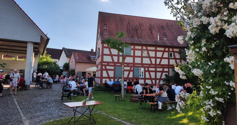 Treffen, Genießen und Bleiben: Begegnungen beim Woi-Eck am BüchnerHaus Tische und Bänke stehen auf dem gepflasterten Hof und dem Rasen vor einem alten Fachwerkhaus - dem Büchnerhaus. An den Tischen sitzen viele Menschen und unterhalten sich. Im Bildvordergrund ist ein weiß blühender Busch zu sehen.