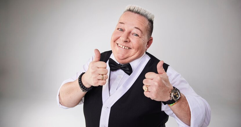 Woody Feldmann kommt nach Riedstadt. Woody Feldmann hat ein weißes Hemd mit schwarzer Fliege und eine schwarze Weste an. Sie streckt beide Daumen nach oben.