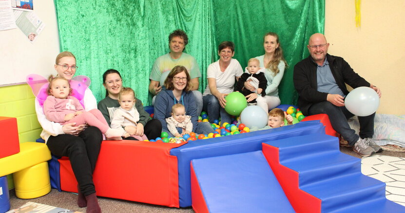 Treffen am Bällebad: Bürgermeister Marcus Kretschmann mit den Gruppenbetreuern Katharina Janzen, Heike Schuhknecht und Mario Quast (hintere Reihe v.re.) sowie Müttern und Kindern der Drop-In-Gruppe. In einem Bällebad mit bunten Bällen sitzen und liegen kleine Kinder. Drumherum sitzen auf der Umrandung Mütter mit ihren Kindern, die Gruppenbetreuer Heike Schuhknecht, Mario Quast und Katharina Jantzen sowie Bürgermeister Marcus Kretschmann.