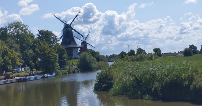 Die Greetsieler Zwillingsmühlen gehören auch zum Besichtigungsprogramm In der Bildmitte ist ein Fluss zu sehen. Links und rechts an seinem Ufer stehen Büsche und Bäume. Links stehen hintereinander zwei gleich aussehende Windmühlen.