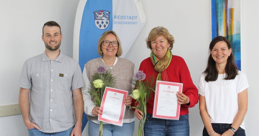 Fachbereichsleiter Rick Hölzer und Fachberaterin Nadja Pfitzner umrahmen die erfolgreichen Absolventinnen Anja Ohl und Anja Fehr (v.li.) Vor einer blau-weißen Fahne mit dem Wappen und der Aufschrift Büchnerstadt Riedstadt stehen Fachberichsleiter Rick Hölzer, die erfolgreichen Absolventinnen Anja Ohl und Anja Fehr sowie Fachberaterin Nadja Pfitzner. Ohl und Fehr halten ihre Zertifikate in den Händen.