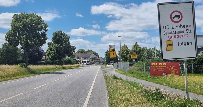 Schilder weisen ausdrücklich auf das Durchfahrtverbot und die Umleitungsstrecke für Lkws hin. Links im Bild ist die Landstraße zu sehen, die durch Leeheim führt, im Hintergrund ist der Ortsanfang. Rechts im Bild zeigen Schilder das Durchfahrtverbot für Lkw und den Pfeil der Umleitungsstrecke.