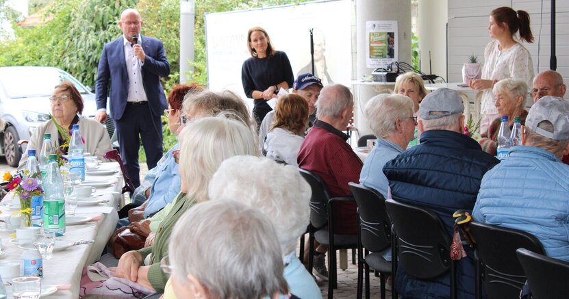 Bildunterschrift: Beste Stimmung herrschte beim Sommerfest der Atempause. Bürgermeister Marcus Kretschmann begrüßte die Gäste, neben ihm steht die Leiterin der Seniorenberatungsstelle Stefanie Drozdzynski. An festlich gedeckten Tischen sitzen ältere Menschen. Vor ihnen steht Bürgermeister Marcus Kretschmann mit Mikrofon und spricht. Neben ihm steht die Leiterin der Seniorenberatungsstelle Stefanie Drozdzynski.