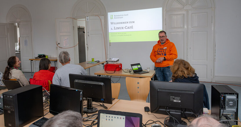 Im Linux-Café gibt es Hilfe zur Selbsthilfe. Von hinten sind Menschen zu sehen, die an Tischen vor ihren aufgeklappten Laptops sitzen. Vorne steht ein Mann und erklärt etwas. An der Wand steht "Willkommen zum 2. Linux-Café"