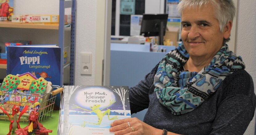 Bei Ellen Stieb in Wolfskehlen können letztmals am 26. März Bücher ausgeliehen werden. Büchereimitarbeiterin Ellen Stieb steht in der Bücherei Wolfskehlen an einem Tisch und zeigt das Bilderbuch "Nur Mut, kleiner Frosch. Auf dem Tisch stehen Tonie-Figuren in Form von Drachen. In einem Drahtkorb befinden sich weitere Kinderbücher.