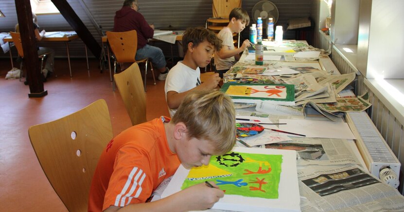 Kreative Konzentration: Manuel, Noah und Lucas (von vorne), im Hintergrund Jugendpfleger Kai Faßnacht und Leia. Ein langer Tisch steht entlang einer Fensterfront. Vorne sitzt ein Junge in orangem T-Shirt und malt an einem Bild mit grünem Hintergrund. Weiter hinten sitzen noch zwei Jungs und malen. Um die Ecke sitzt Jugendpfleger Kai Faßnacht mit dem Rücken zum Betrachter und malt auch.