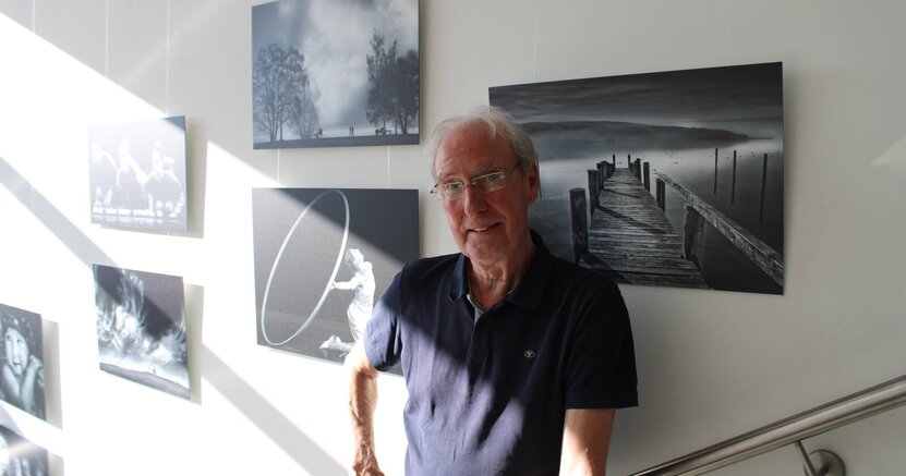 Bernd Susenburger vor einigen seiner Werken in der Treppenhausgalerie. Bernd Susenburger steht vor einigen seiner Schwarz-Weiß-Fotografien, die an Wänden des Treppenhauses im Rathaus hängen. Das Bild rechts von ihm zeigt einen Steg, der ins Wasser eines Sees führt, links ist unter anderem ein Bild von einer knienden Frau zu sehen, die mit ihren Armen hinter sich einen großen Reif hält.