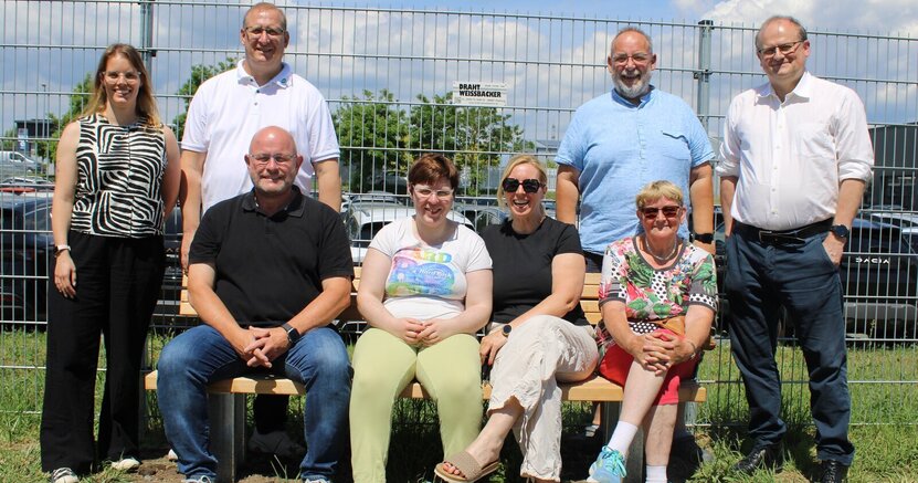 Auf der neuen Bank freuen sich Bürgermeister Marcus Kretschmann, Katja Glock, Ilona Jahnke und Ursula Fraikin (v.li.), stehend Nachhaltigkeitsmanagerin Sandra Seum, Mathias Lachmann, Gaston Wolfsturm und Michael Fraikin (v.li.) über di Auf der neuen Bank sitzenBürgermeister Marcus Kretschmann, Katja Glock, Ilona Jahnke und Ursula Fraikin (v.li.), daneben und dahinter stehen Sandra Seum, Mathias Lachmann, Gaston Wolfsturm und Michael Fraikin (v.li.). Im Hintergrund sind ein Zaun und dahinter Autos zu sehen.