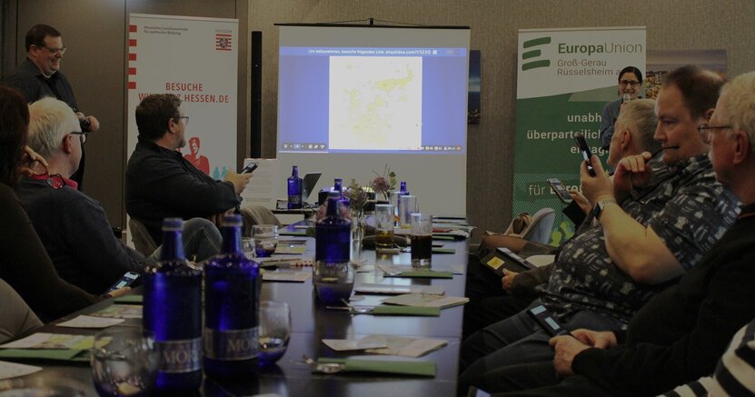 Andreas Christ und Dr. Birgit Bujard führten durch den Abend. An einem langen Tisch sitzen Menschen mit ihren Smartphones. Auf einer Leinwand ist eine Frage des Pub Quiz zu sehen. Links und rechts davon stehen die Moderatoren Andreas Christ und Dr. Birgit Bujard mit Mikrofonen in der Hand.