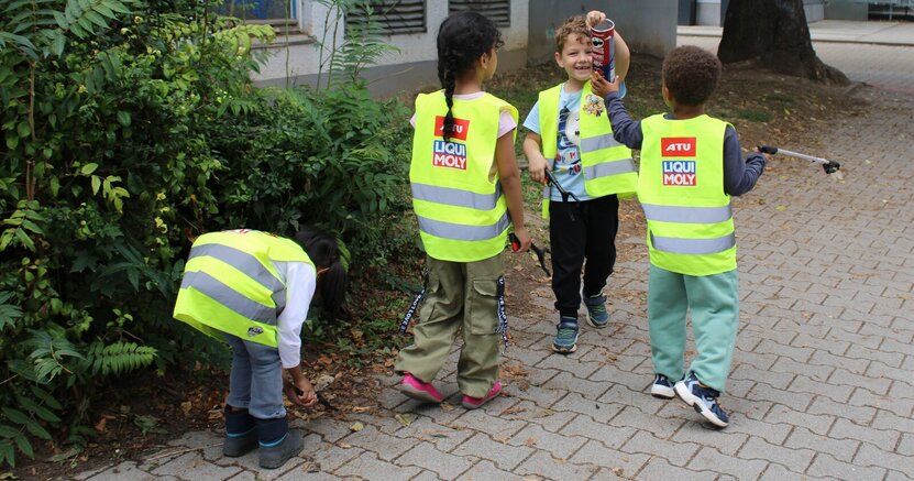 Auch eine Chips-Dose gehörte zu den Funden der Kitakinder Mehrere Kindergartenkinder haben gelbe Warnwesten an und stehen am Rande eines Fußwegs. Ein Junge hält eine Chipsdose hoch. Ein anderes Kind bückt sich gerade und ein weiteres hält eine Greifzange in der Hand.