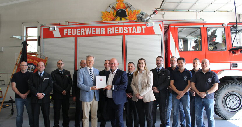 Innenstaatssekretär Martin Rößler übergab Bürgermeister Marcus Kretschmann den Förderbescheid In der Fahrzeughalle der Freiwilligen Feuerwehr Erfelden stehen in einer langen Reihe vor einem Feuerwehrwagen mit der Aufschrift "Feuerwehr Riedstadt" Feuerwehrleute in Uniform. Vor ihnen überreicht Innenstaatsssekretär Martin Rößler den Zuwendungsbescheid an Bürgermeister Marcus Kretschmann.