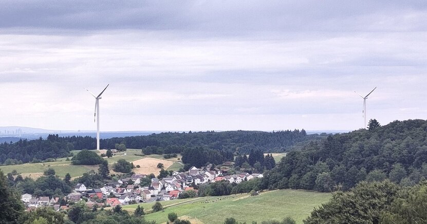 Die Windkraft ist ein wichtiger Baustein der Energiewende. In einiger Entfernung von Wiesen und Wäldern sowie einer Siedlung sind zwei Windkrafträder zu sehen.