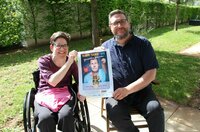 Die Behindertenbeauftragte Marina Wohner sitzt im Rollstuhl. Neben ihr sitzt Kulturbüroleiter Marco Hardy auf einem Stuhl. Gemeinsam halten sie ein Plakat mit der Veranstaltung von Martin Fromme hoch.