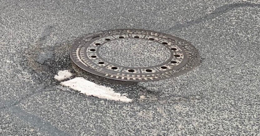 Schachtabdeckungen müssen saniert werden. Abgebildet ist eine runde Schachtabdeckung mit einer Umrandung mit Löchern aus Metall. Sie befindet sich inmitten einer Straße mit Asphaltdecke.