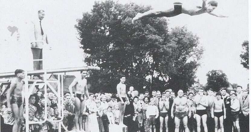 Mit einem Sprung ins Schwimmerbecken eröffnete Peter Bär 1956 das Freibad Goddelau. Hinter ihm steht sein Vater, Bürgermeister Christoph Bär. Auf der schwarz-weiß-Fotografie stehen Menschen in Badesachen um ein Schwimmbecken. Vom Sprungbrett macht ein junger Mann einen Kopfsprung. Dahinter steht ein Mann im Anzug.
