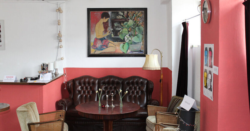 Im gemütlichen Theatercafé tagt der "Club der Dichter". Im Theatercafé der BüchnerBühne steht an der Wand ein gemütliches Sofa, davor ein runder Tisch und drumherum einige Sessel. An der Wand hängt ein großes Gemälde.