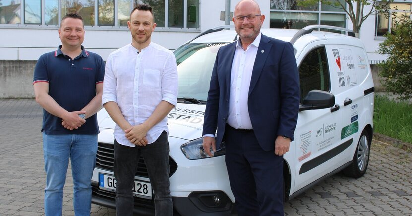 Über das Auto freuen sich Marcus Schramm und Christian Pudel vom IT-Service sowie Bürgermeister Marcus Kretschmann (v.li.) Ein weißes Auto mit verschiedenen Aufschriften steht auf einem Parkplatz hinter dem Riedstädter Rathaus. Auf der Motorhaube ist der Schriftzug "Büchnerstadt Riedstadt" zu erkennen. Vor dem Wagen stehen Marcus Schramm und Christian Pudel von der IT-Abteilung sowie Bürgermeister Marcus Kretschmann