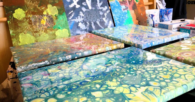 Gemälde, die in einem der Kunstworkshops entstanden sind Bunte, glänzende Bilder in Mamoriertechnik liegen auf einem Tisch aus. Dahinter lehnen Bilder an einer Stütze. Bei genauem Hinsehen sieht man, dass sich in der Mitte ein Foto befindet. Das Motiv darauf wurde auf dem Bild weitergemalt.
