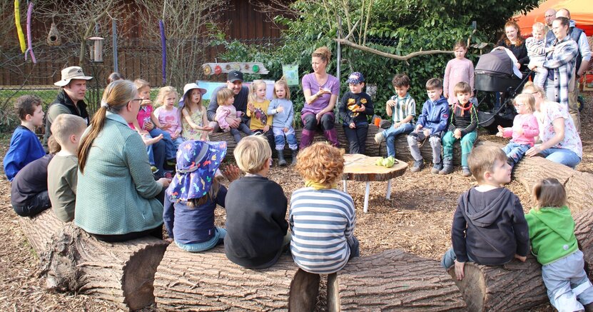 Die Kinder des Naturkindergartens sangen mit Kitaleiterin Anja Llorca (Mitte) ein Begrüßungslied. Die Kinder des Naturkindergartens sitzen mit ihren Betreuern auf querliegenden Baumstämmen, die zu einem Rund gelegt wurden. Sie klatschen und singen.