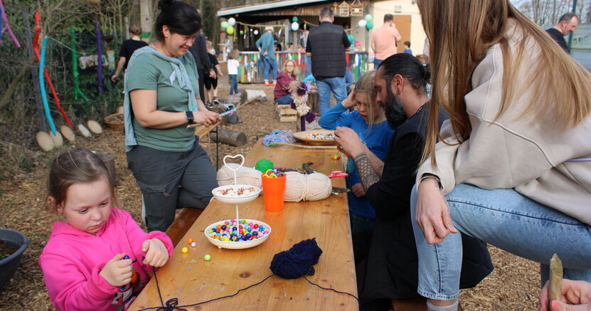 Bastelangebot beim "Tag des Waldes" im letzten Jahr. An einem langen Tisch draußen basteln Kinder und Erwachsene mit Wollfäden und Perlen. Im Hintergrund sind Bäume zu sehen.