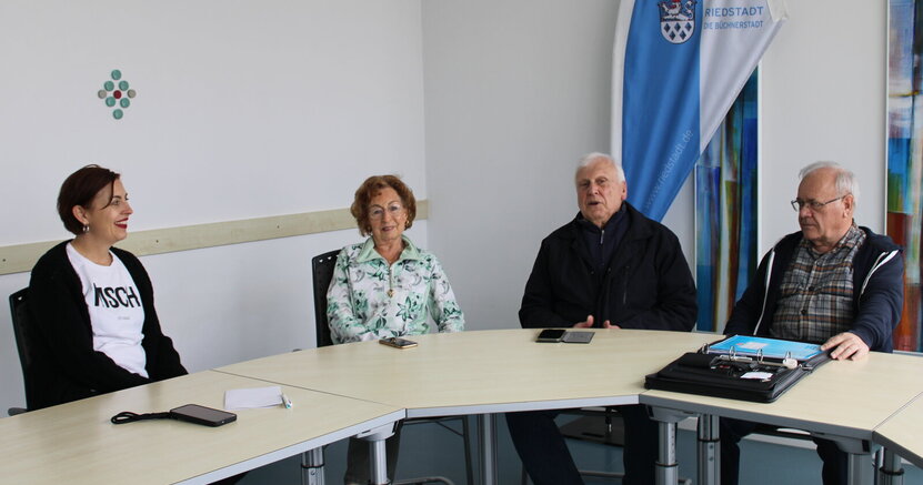 Gleichstellungsbeauftragte Jennifer Muth, Brigitte Lautenbach, Heinz Osterod und Berater Hans-Jürgen Pretzel in einem Raum des Riedstädter Rathauses. Gleichstellungsbeauftragte Jennifer Muth, Brigitte Lautenbach, Heinz Osterod und Berater Hans-Jürgen Pretzel sitzen im Halbrund an einem langen Tisch, im Hintergrund ist die Fahne der Büchnerstadt Riedstadt zu sehen. Sie ist vertikal in eine blaue und eine weiße Seite unterteilt und versehen mit dem Stadtwappen und der Aufschrift in Blau "Büchnerstadt Riedstadt".