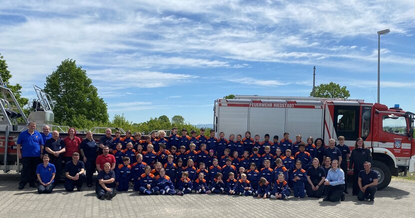 64 stolze Feuerwehrkinder mit ihren Ausbildern und Betreuern und der Stadtjugendfeuerwehrwartin Natascha Spalke (hellblaues Hemd) Kinder der Kinderfeuerwehren Riedstadt stehen mit ihren blau-orangen Uniformen vor einem Feuerwehrauto. Sie werden umringt von erwachsenen Betreuern.