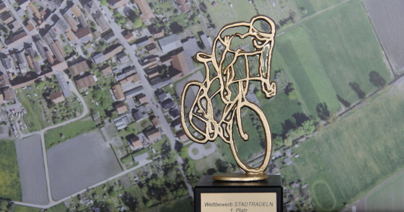 Die Büchnerstadt möchte den Wanderpokal als fahrradaktivste Kommune im Kreis Groß-Gerau möglichst in Riedstadt behalten. Zu sehen ist der Wanderpokal in Form eines Radfahrers. Im Hintergrund ist ein Foto von oben von Straßen und Feldern zu sehen.