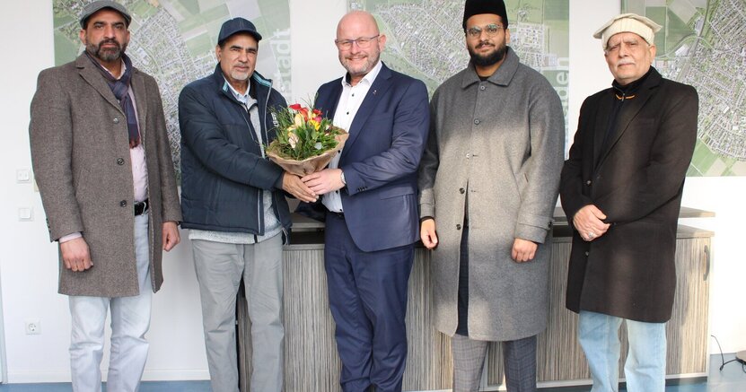 Atta-ul Ghafoor, Abdul Shakoor, Bürgermeister Marcus Kretschmann, Imam Awayes Ahmad Malik und Mumtaz Ahmad (v.li.) im Arbeitszimmer des Bürgermeisters. Atta-ul Ghafoor, Abdul Shakoor, Bürgermeister Marcus Kretschmann, Imam Awayes Ahmad Malik und Mumtaz Ahmad (v.li.)stehen im Arbeitszimmer des Bürgermeisters vor Wandbildern, die Luftaufnahmen der Stadtteile zeigen. Der Bürgermeister hat einen Blumenstrauß in der Hand.