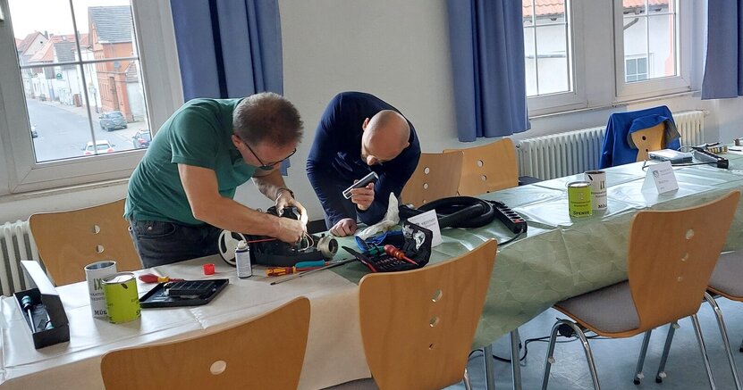Gemeinschaftlich gewerkelt wird beim Reparatur-Café im alten Rathaus Crumstadt. Zwei Männer beugen sich an einem Tisch über ein elektrisches Gerät. Der eine Mann leuchtet mit einer Taschenlampe, während der andere versucht, das Gerät zu reparieren.