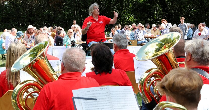 Der Musikverein Goddelau mit seinem Dirigenten Thomas Grimm begeisterte beim Picknick-Konzert im Volkspark. Im Vordergrund sind Musiker des Musikvereins in roten T-Shirts zu sehen. Sie sitzen mit dem Rücken zum Betrachter. Vor ihnen steht Dirigent Thomas Grimm. Im Hintergrund sind Zuschauer auf Bierbänken im Volkspark Goddelau zu sehen. Hinter ihnen stehen Bäume.