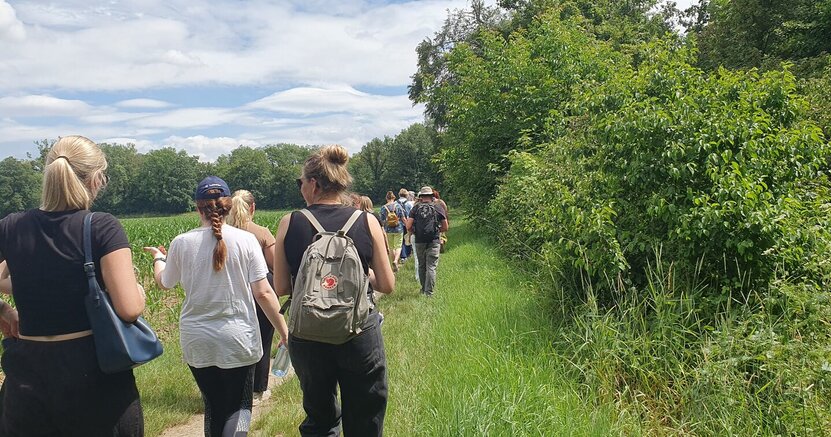 Auch eine Kräuterwanderung gehörte zu den Angeboten Von hinten sind Frauen in Sommerkleidung zu sehen, die einen Wiesenweg entlang gehen. Auf der linken Seite befinden sich Felder, auf der anderen Seite Büsche und Bäume.