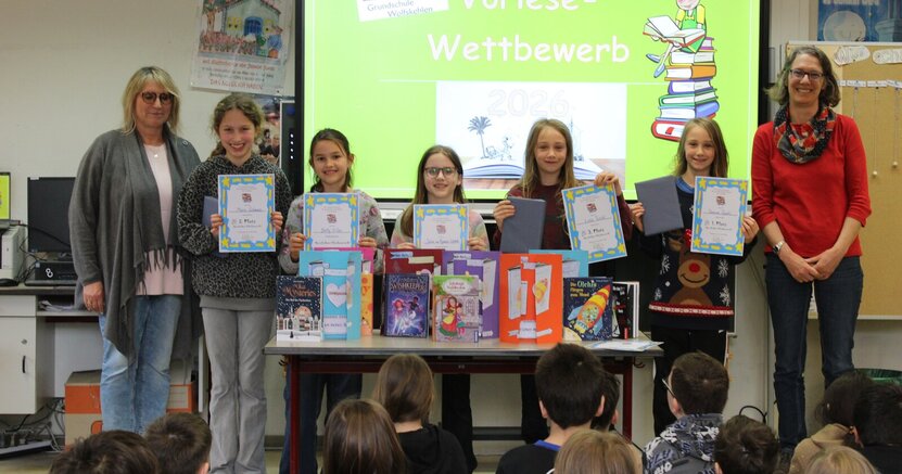 Schulleiterin Susanne Möller mit Marie, Betty, Julia, Luise, der Erstplatzierten Theresa und Büchereileiterin Anja Stark (v.li.). Schulleiterin Susanne Möller steht mit Marie, Betty, Julia, Luise, der Erstplatzierten Theresa und Büchereileiterin Anja Stark (v.li.)vor den auf dem Boden sitzenden Viertklässlern. Die Vorlese-Kinder halten ihre Urkunden hoch. Im Hintergrund ist auf einer Leinwand zu lesen: "Vorlesewettbewerb 2026"