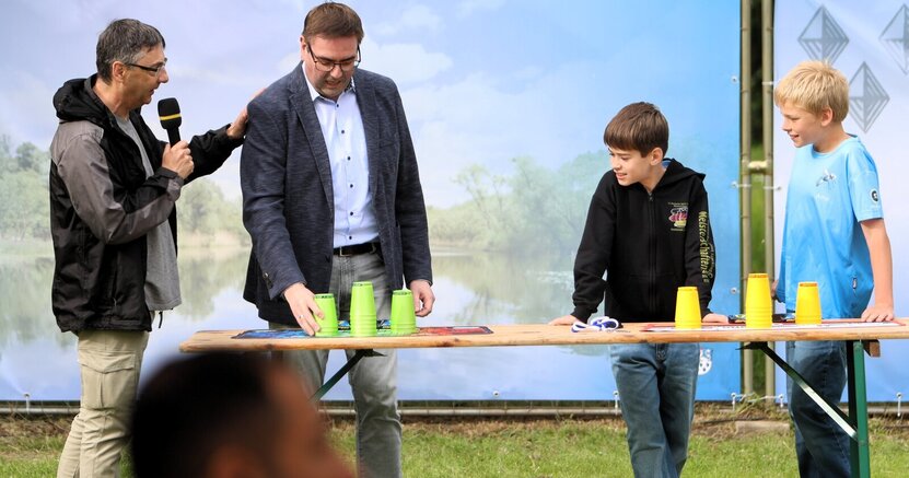 Stadtverordnetenvorsteher Guido Funk probierte sich auch im Speed Stacking. Trainer Robert Sinkovic (links) erklärte unter den Blicken der Jung-Profis Ben Bittner und Arne Rupf die Regeln. Vor dem Publikum der Jugendsportlerehrung steht auf der Wiese im Freibad Goddelau ein Tisch mit grünen und gelben Bechern für Speed Stacking. Stadverordnetenvorsteher Guido Funk greif gerade nach den grünen Bechern, um sich im Becherstapeln zu versuchen. Neben ihm steht Trainer Robert Sinkovic mit Mikrofon, um die Regeln zu erklären. Auf der anderen Seite von Funk stehen die Jugendlichen Ben Bittner und Arne Rupf, die danach ihr Können zeigen werden.